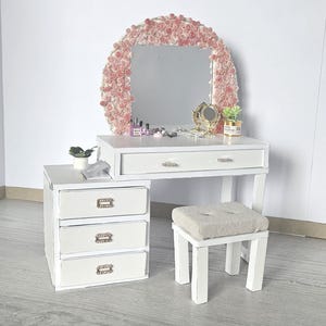May include: A white vanity set with a matching stool. The vanity has a mirror with a pink floral frame, a drawer, and a side cabinet with three drawers. A small plant and accessories are on the vanity.