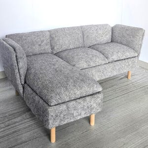 May include: A gray sectional sofa with a chaise lounge. The sofa has a textured fabric and wooden legs. The chaise lounge is on the left side. The sofa is a neutral color and would fit in many home decor styles.