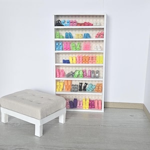 May include: A white miniature shoe shelf filled with colorful doll shoes. The shelf is next to a small, upholstered ottoman with a white base. The shoes are arranged by color, including pink, yellow, and blue.