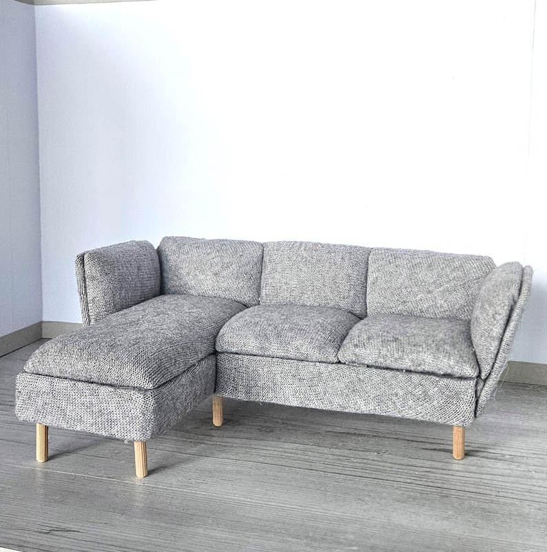 May include: A gray sectional sofa with a chaise lounge on the left side. The sofa has a textured fabric and light-colored wooden legs. The design is modern and minimalist, suitable for a dollhouse or miniature setting.