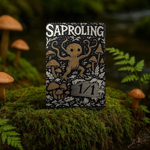 Op de afbeelding: Een zwart-wit kaart met het woord "SAPROLING" bovenaan. De kaart toont een gouden illustratie van een wezen met paddenstoel- en plantdetails. Het nummer "1/1" staat onderaan. De kaart bevindt zich in een mosachtig bos.