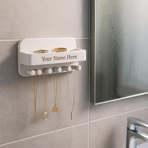 Könnte beinhalten: Weißer, an der Wand befestigter Schmuck-Organizer mit Haken und einem Regal. Das Regal enthält Ringe, und die Haken zeigen Halsketten. Der Text "Your Name Here" ist auf dem Regal aufgedruckt. Der Schmuck ist gold- und silberfarben.