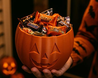 Puede incluir: Un cuenco naranja con forma de calabaza lleno de varios caramelos envueltos. El cuenco tiene una cara de linterna tallada. La persona que sostiene el cuenco lleva un suéter naranja con un diseño de murciélago. Dos calabazas iluminadas están en el fondo.