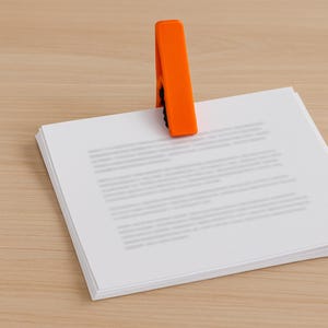 May include: An orange plastic clip holding a stack of white paper. The clip is positioned at the top of the paper, securing the pages together. The background is a light brown wooden surface.