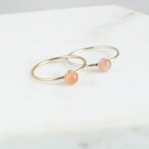 May include: Two delicate rings with a peach-colored gemstone. The ring on the left is gold, and the ring on the right is silver.