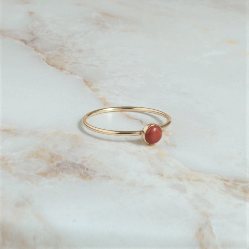 Puede incluir: Un anillo de oro simple con una peque&ntilde;a piedra redonda de color rojo.