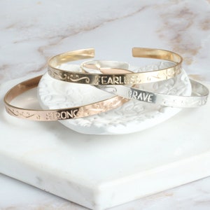 May include: Three gold, silver, and rose gold bangle bracelets with engraved words "Strong", "Fearless", and "Brave".