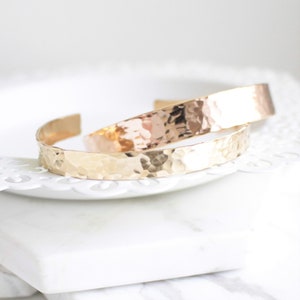 May include: Two hammered metal bracelets, one gold and one rose gold, on a white surface.