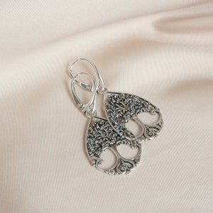 May include: A pair of silver-toned drop earrings. Each earring features a detailed tree design within a teardrop shape. The earrings have a lever-back closure and are displayed on a light pink fabric.