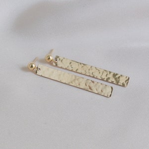 May include: A pair of gold bar earrings with a hammered texture. The earrings have a simple, minimalist design.