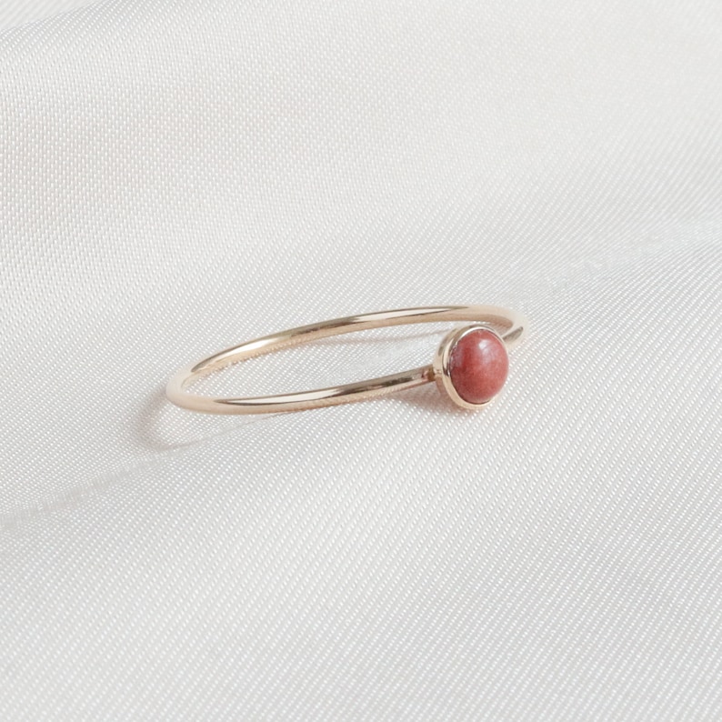 Puede incluir: Un delicado anillo de oro con una sola piedra redonda de color rojo.