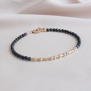 May include: Delicate bracelet featuring small, dark blue beads and gold-colored accents. The bracelet has a clasp closure and a section of gold beads and bars. The jewelry is displayed on a light pink surface.