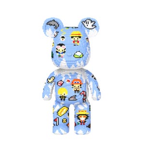 May include: A light blue bear-shaped figurine with pixelated details. The figurine is decorated with various cartoon characters and symbols in multiple colors. The bear has large ears and is standing upright.