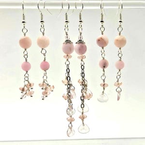 May include: A collection of six dangle earrings. Each earring features a silver hook, a large pink bead, a smaller pink bead, and dangling pink and clear crystal accents. The earrings are displayed against a white background.