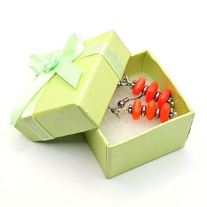 May include: A pair of drop earrings featuring bright orange oval beads and silver-toned metal spacers, presented inside a light green square cardboard gift box with a matching ribbon bow on the lid and white foam padding inside.