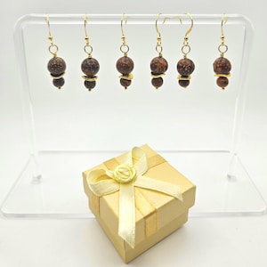 May include: Six pairs of dangle earrings with brown and gold beads are displayed on a clear stand. Each earring has a gold hook and a combination of round and oval beads. A small, gold gift box with a cream ribbon and bow sits in front.