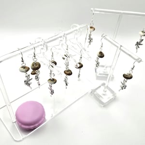 May include: A display of handmade earrings with a natural, earthy aesthetic. The earrings feature a combination of silver-toned metal and round, speckled shell-like beads. A light purple macaron sits on the display stand.