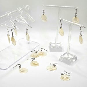 May include: Earrings with a layered, circular design. The earrings are a light, off-white color and hang from dark metal hooks. Some earrings are displayed on clear acrylic stands, while others are arranged on a white surface. The earrings have a natural, organic look.