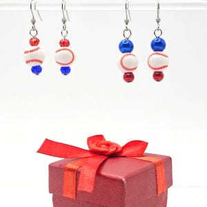 May include: Two pairs of dangling earrings featuring white baseball beads with red stitching. The first pair has red and blue faceted beads, while the second pair has blue and red smooth round beads. All earrings hang from silver-toned metal hooks.