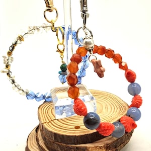May include: Two beaded bracelets with keychains. One bracelet features clear and gold beads, the other has orange, red, and blue beads. The bracelets are displayed on a wooden slice and clear acrylic stands.
