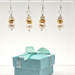 May include: Four pairs of dangle earrings with white pearl drops, gold-colored metal accents, and clear beads. The earrings hang from silver-colored hooks. A light blue gift box with a ribbon is in the foreground.