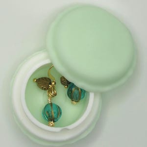 May include: A pair of teal and gold earrings inside a light green, round jewelry box. The earrings feature teal beads and gold-colored leaf accents. The box is open, revealing the earrings against a matching green interior.