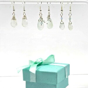 May include: A collection of six pairs of silver earrings with clear, teardrop-shaped gemstones. The earrings hang from a clear bar. A teal gift box with a white ribbon and bow is in the foreground.