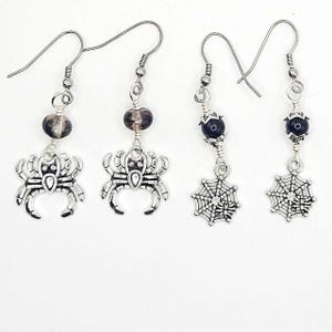 May include: Two pairs of silver-toned dangle earrings. The first pair features a spider charm with a translucent grey bead. The second pair features a spiderweb charm with a small spider and a dark blue bead. All earrings have silver-toned hooks.