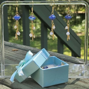 May include: Four pairs of dangle earrings with blue and white beads, displayed on a clear stand. The earrings have gold-colored hooks and chains. A light blue gift box with a ribbon is open, revealing more jewelry.