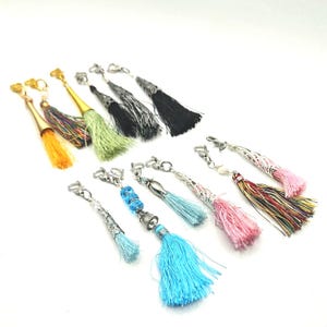 May include: A collection of colorful tassel keychains. The tassels come in various colors, including yellow, green, black, blue, and pink. Each keychain has a metal clasp and decorative top. The tassels are made of a soft, silky material.