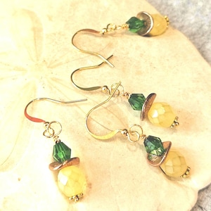 May include: Three dangle earrings featuring gold-tone fishhook wires, each with a faceted green crystal bead, a small copper-colored metal disc, and a pale yellow faceted bead at the base. The earrings are arranged in a cluster on a light surface.