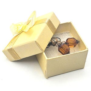 May include: A pair of amber-colored faceted glass bead earrings with dark metal accents, resting inside a square, light yellow cardboard gift box that features a matching yellow ribbon bow on the lid and a white foam insert base.
