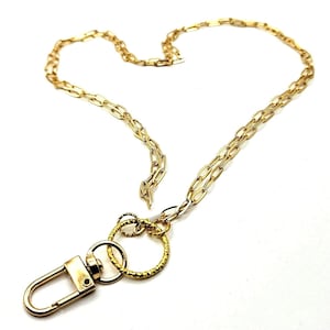 May include: A gold-toned metal lanyard featuring a long chain with elongated oval links. Attached to the end of the chain is a circular ring with a textured surface, connected to a smaller smooth ring and a rectangular lobster claw clasp.