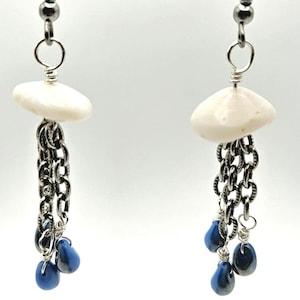 May include: A pair of handmade earrings featuring a white, disc-shaped top, a silver chain, and three blue teardrop-shaped beads. The earrings have a unique, dangling design, perfect for adding a touch of elegance to any outfit.
