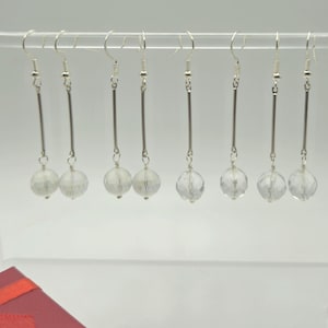 May include: A collection of silver-toned dangle earrings. Each earring features a long, thin bar suspended from a hook, with a faceted, clear crystal ball at the bottom. The earrings are displayed on a clear, curved stand.