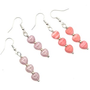 May include: Three pairs of dangle earrings with heart-shaped beads. Two pairs feature pink beads, while the third pair has lavender beads. Each earring has three heart beads strung together with silver-colored metal accents and ear wires.