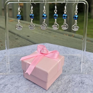 May include: Six pairs of dangle earrings with blue faceted beads and silver-tone crescent moon and floral charms are displayed on a clear stand. A small pink gift box with a matching pink ribbon and bow sits in front of the earrings.