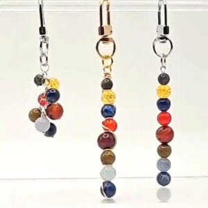 May include: Three decorative charms featuring multi-colored spherical beads in shades of yellow, blue, red, brown, and grey. The left charm has a cluster of beads, while the center and right charms feature vertical strands of beads on metal hooks.