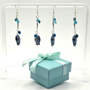 May include: Four pairs of dangle earrings with silver hooks. Each earring features a dark blue, oval-shaped stone, accented with small blue beads and silver wire. A light blue gift box with a matching ribbon and bow is in the foreground.