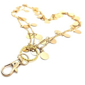 May include: A gold-toned lanyard featuring a delicate chain adorned with numerous small, flat, circular discs. The accessory includes a textured gold ring connected to a functional lobster claw clasp, designed for holding keys or identification badges.