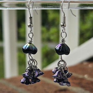 May include: A pair of dangle earrings with iridescent dark blue and purple beads. The earrings have a silver-tone metal hook and wire design. The top bead is a teardrop shape, and the bottom has a cluster of small flower-shaped beads.