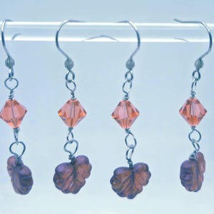May include: Four pairs of dangle earrings. Each earring features a silver-tone hook, a peach-colored, diamond-shaped bead, and a dark purple leaf-shaped charm. The earrings are suspended from a clear bar.
