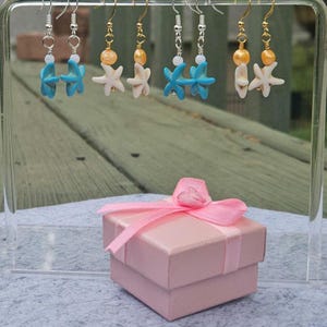 May include: A display of six pairs of earrings with starfish and bead accents. The earrings are in various colors, including turquoise, white, and gold. A pink gift box with a matching ribbon is in the foreground.