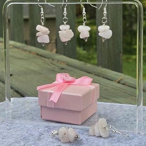 May include: Four pairs of rose quartz earrings with silver-colored hooks are displayed. A pink gift box with a matching ribbon sits below. One pair of earrings rests on the surface. The earrings feature irregular, light pink stones.