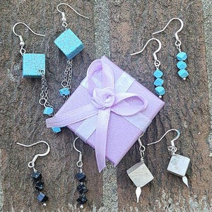 May include: An assortment of handmade earrings in various designs. The earrings feature turquoise, black, and gray square and cube-shaped beads, and silver hooks. A small, lavender gift box with a white ribbon is also present.