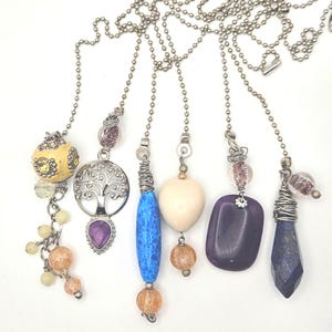 May include: A collection of decorative ceiling fan pull chains with various pendant designs. The pendants include a yellow orb, a tree of life, a blue stone, a cream heart, a purple stone, and a dark blue crystal. Each chain is silver-toned.