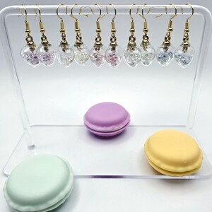 May include: A display of handmade earrings with gold-colored hooks and clear, heart-shaped pendants filled with colorful glitter. Three macarons in pastel colors are also displayed: lavender, mint green, and yellow.