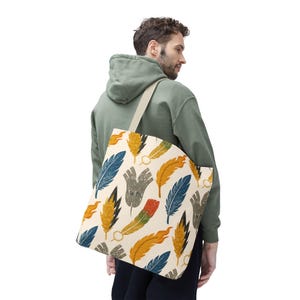 May include: A cream-colored tote bag featuring a repeating pattern of colorful feathers in shades of blue, yellow, orange, and gray. The bag has a beige strap and is carried over the shoulder.
