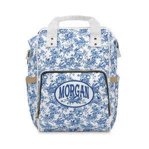 May include: A white and blue floral print backpack with the name "MORGAN" in a blue oval on the front. The backpack has a top handle, adjustable straps, and multiple pockets. The floral pattern features rabbits and other elements.