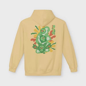 May include: A pale yellow hoodie with a green and red graphic on the back. The graphic features three cartoon characters with Japanese text. The hoodie has a hood and long sleeves.